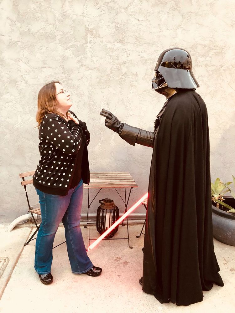 Me (woman with brown hair, wearing blue jeans, black clogs, and a Star Wars cardigan) standing in profile with hands to my throat while a Darth Vader cosplayer carrying a red lightsaber has his hand out in the "force choke" position. Image from a few years ago.