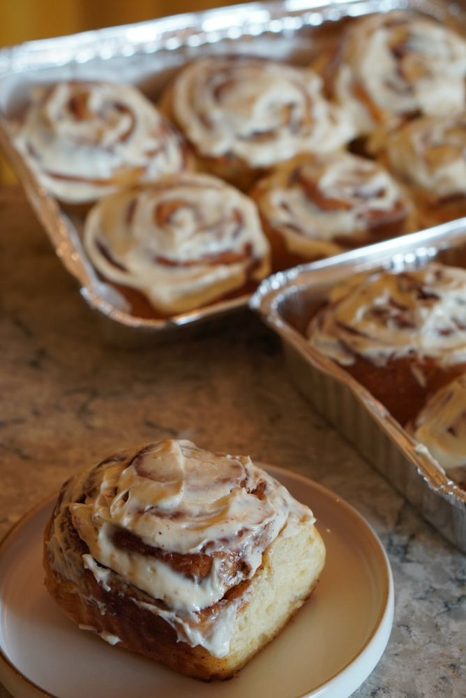 Cinnamon rolls de longa fermentação feitos por mim. Em primeiro plano está um roll em um prato de cerâmica e ao fundo duas bandejas de cinnamon rolls assados. Todos são cobertos com calda de cream cheese com baunilha e açúcar. 