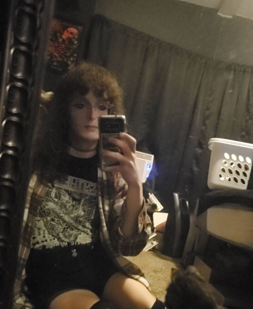 photo of a young woman wearing a grunge style outfit with a shirt from a metal band taking a selfie in a mirror while sitting on the ground.