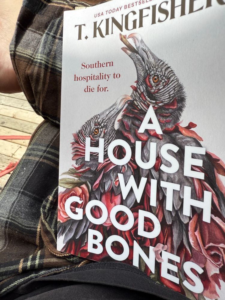 Photo of the book cover of A House with Good Bones by T. Kingfisher on my lap as I sit on the back deck.