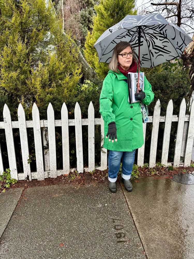 Woman wearing eye glasses, green rain coat, red scarf,  fingerless gloves jeans, and boots holding an umbrella and cradling a book in one arm points to a "1907" sidewalk date stamp. Some bushes and a white picket fence and behind her.  Photo by Linda Yip 