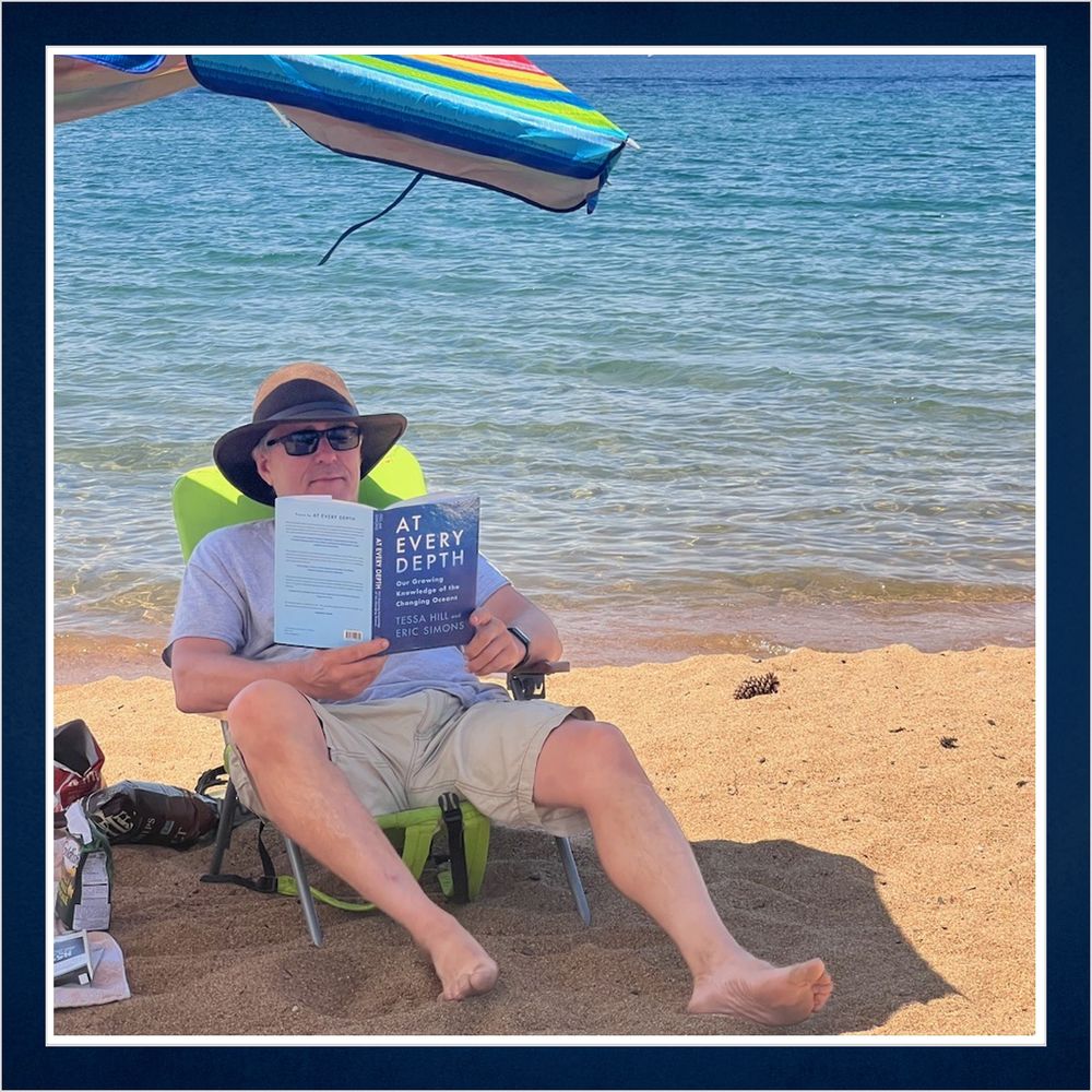 A person sits on a beach under an umbrella with a copy of At Every Depth. Photo by J. Foley