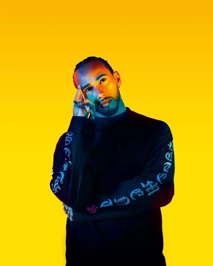picture of lewis hamilton in a black sweatshirt, against a yellow background. he is pictured from the waist up, with one arm crossed across his chest and the other holding his head up. he looks cute as fuck
