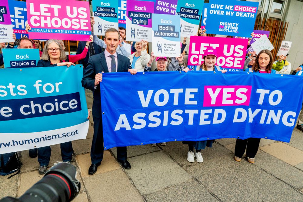A group of demonstrators holding signs advocating for assisted dying. The main blue banner reads "VOTE YES TO ASSISTED DYING." Other signs include "BACK US CHOOSE," "CHOICE & COMPASSION ASSISTED DYING," and "LET US CHOOSE." One sign on the left reads "PROTESTS FOR CHOICE HUMANISM.SCOT." The people are standing on a pavement with a camera in the foreground.
