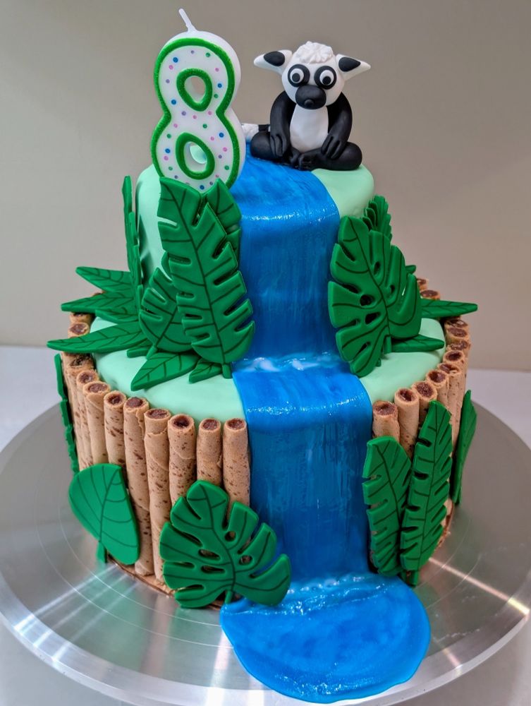 two-tier jungle cake with fondant lemur and #8 candle on top, fondant waterfall through center, and fondant leaves all around. Rolled wafer cookies surround bottom layer suggesting a bamboo fence