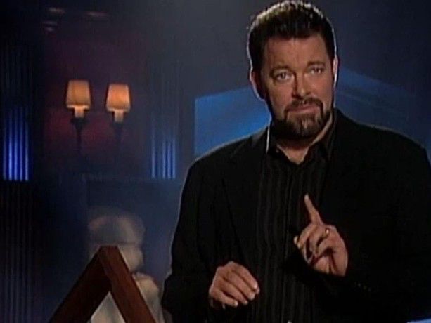 Big Johnny F standing on a dark set with one index finger raised like he’s telling you to wait for something (not clear what, possibly he’s about to swing his leg over the back of a chair) 