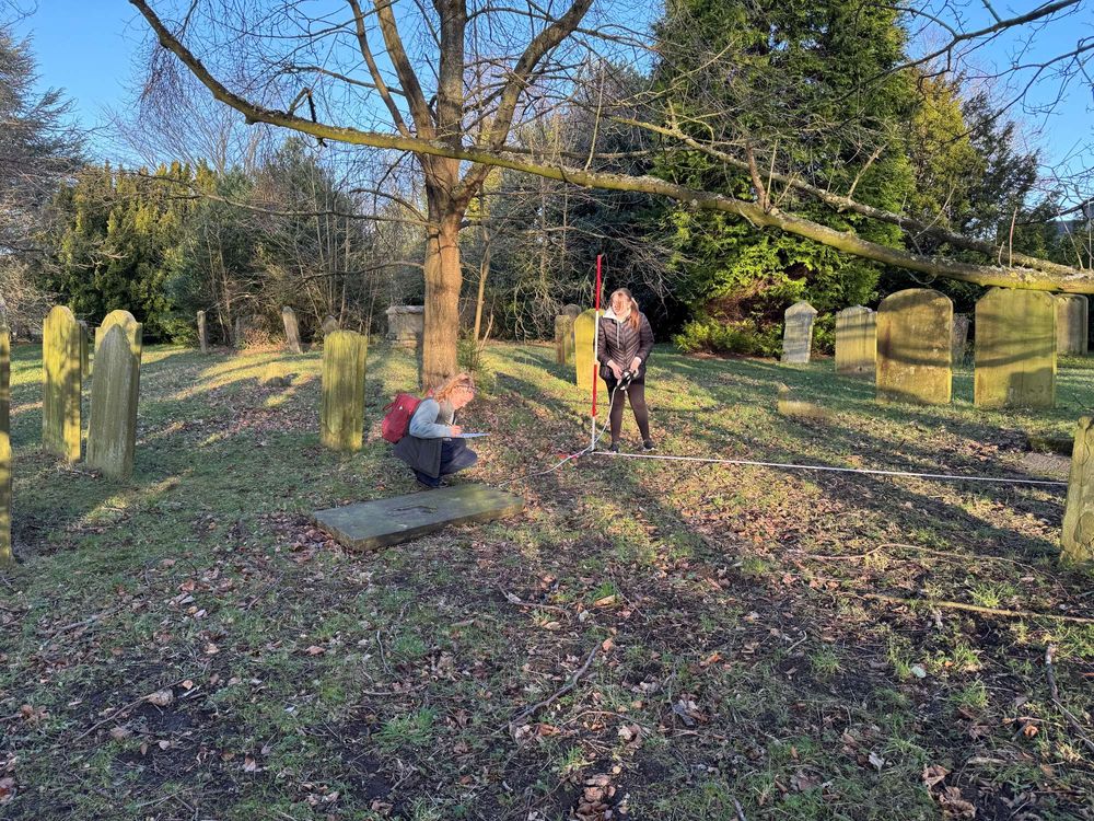 Two students in St Oswald's Churchyard documenting grave monuments using surveyor's tape measure and a ranging pole.