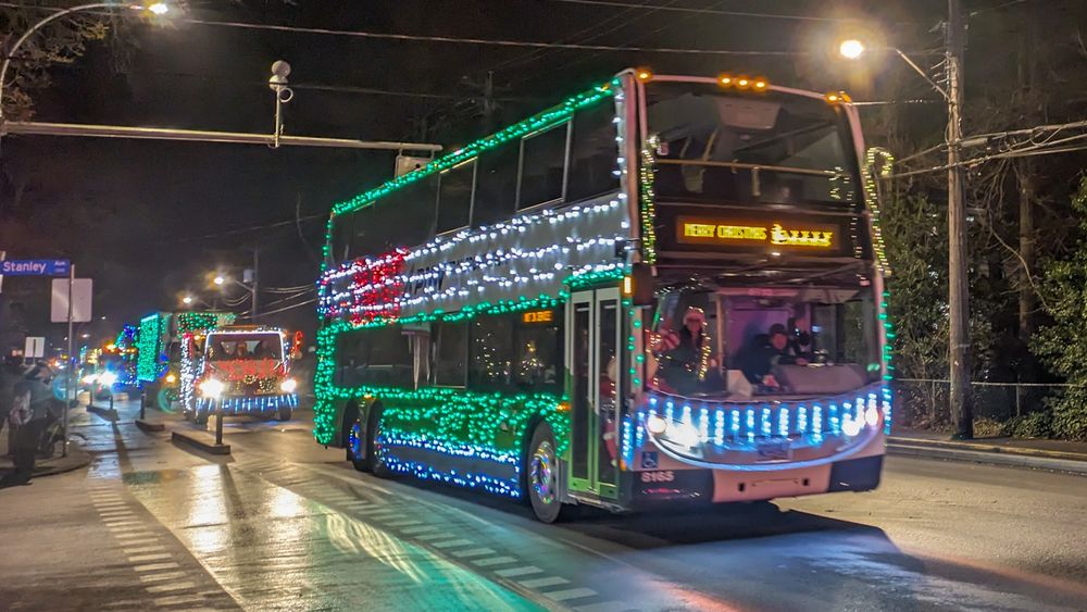 A double decker bus covered in green and white Christmas lights in a parade last weekend. The bus is heading Westbound beside a protected bike lane from the intersection of Fort Street and Stanley Avenue in Victoria, BC.