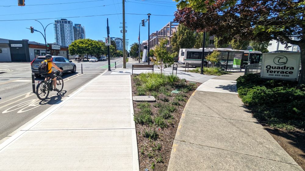The upgraded intersection (Caledonia Avenue and Quadra Street) after the slip lane was removed. This upgrade improved walk & roll safety and added seating, bike racks, a bus shelter, plants and a couple of new trees. The Caledonia Ave protected bike lane is seen on the left.