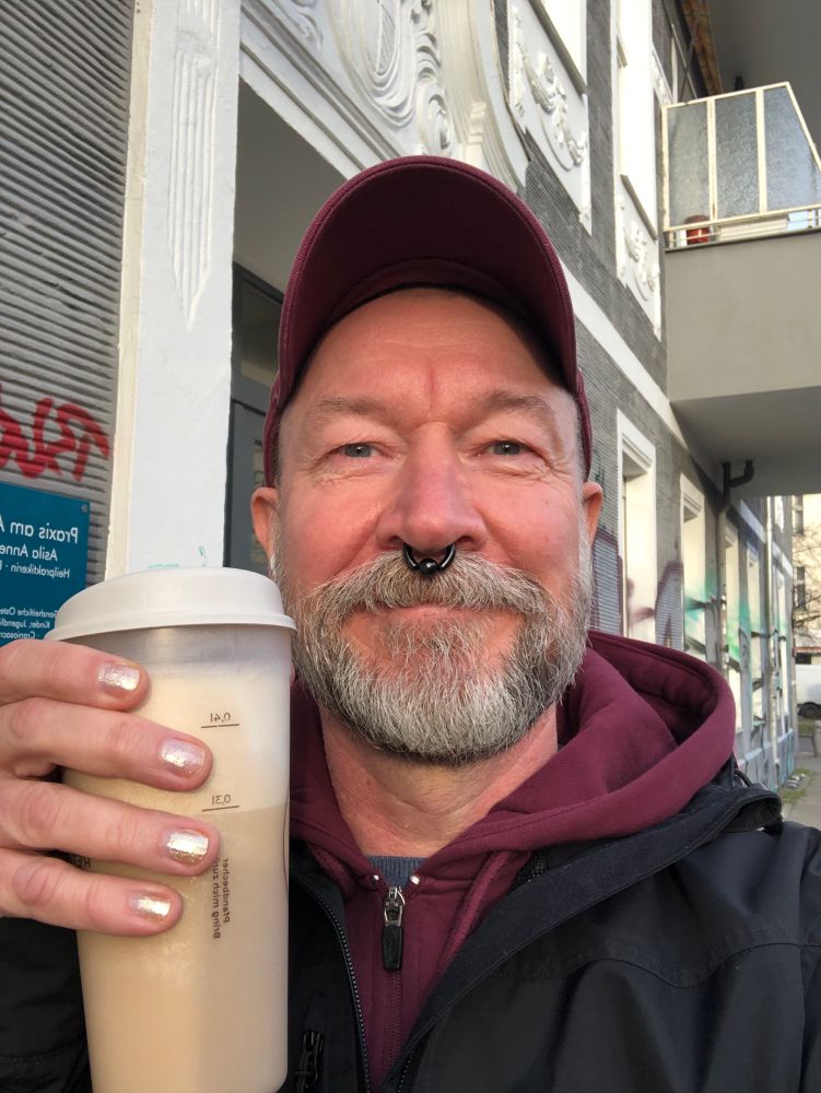 frank steht im prenzlauer berg vor einem haus und hält lächelnd einen kaffee in einem coffee-to-go-becher in die kamera.