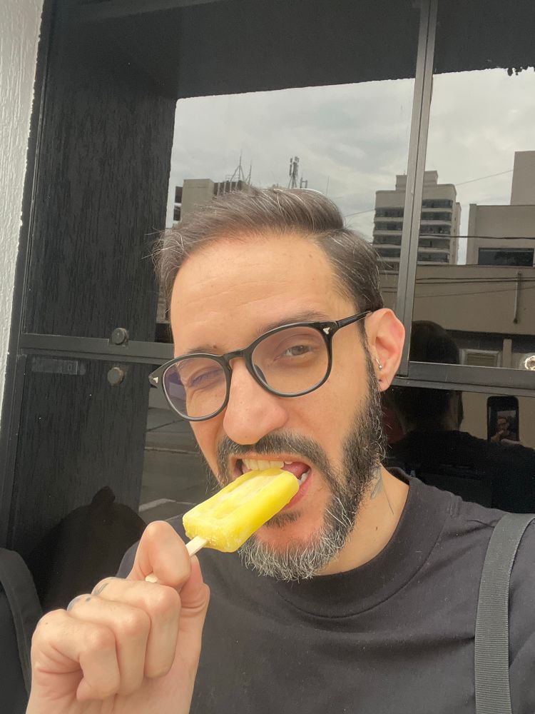 Homem latino de barba grisalha no queixo, de óculos de grau arredondado, camiseta preta dando uma mordida em um picolé amarelo de abacaxi e dando uma piscadela com o olho direito.