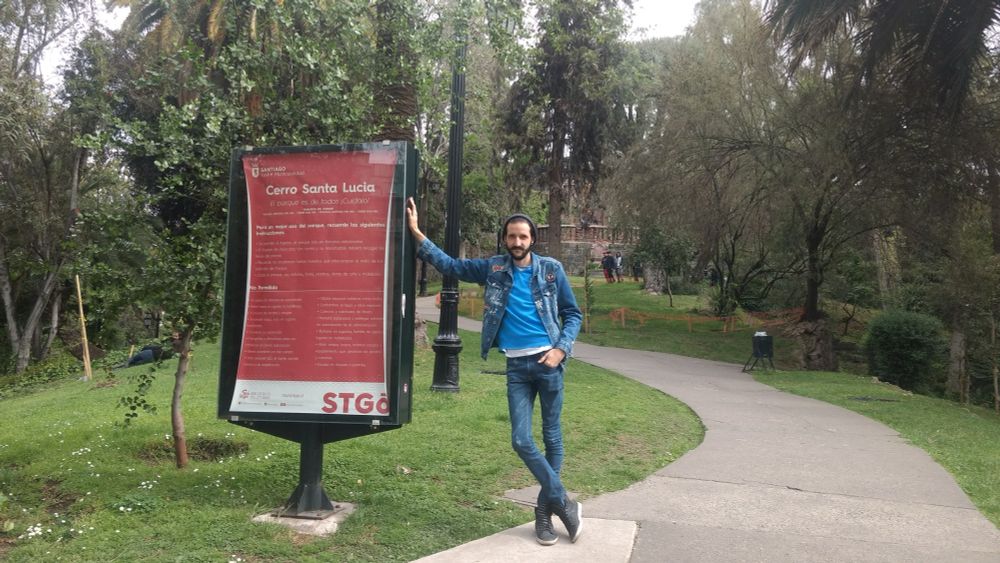 Eu posando ao lado de uma placa do Cerro Santa Lúcia em Santiago de Chile. Com o parque ao fundo com árvores e um caminho de pavimento.