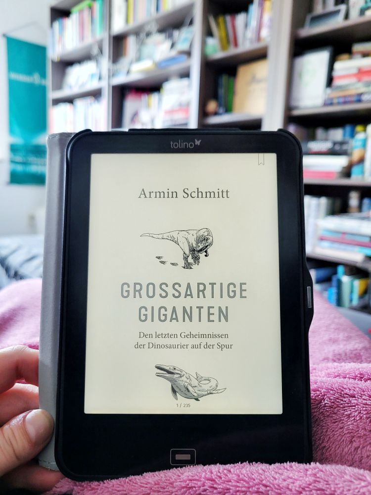 Ich halte einen e-Reader in der Hand. Darauf ist das Buch "Großartige Giganten. Den letzten Geheimnissen der Dinosaurier auf der Spur" von Armin Schmitt geöffnet. Im Hintergrund sieht man meine Bücherregale. 