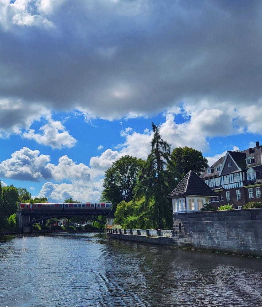 Blick auf die Alster. Im Hintergrund ist eine Brücke, über die eine U-Bahn fährt. Rechts im Bild ist eine Mauer, darauf eine Art kleine Kuppel mit Dach und Fenstern. Dahinter ein Haus.
Der Himmel ist bewölkt, unten eher kleine weiße Wolken, oben im Bild eine große graue Wolke.