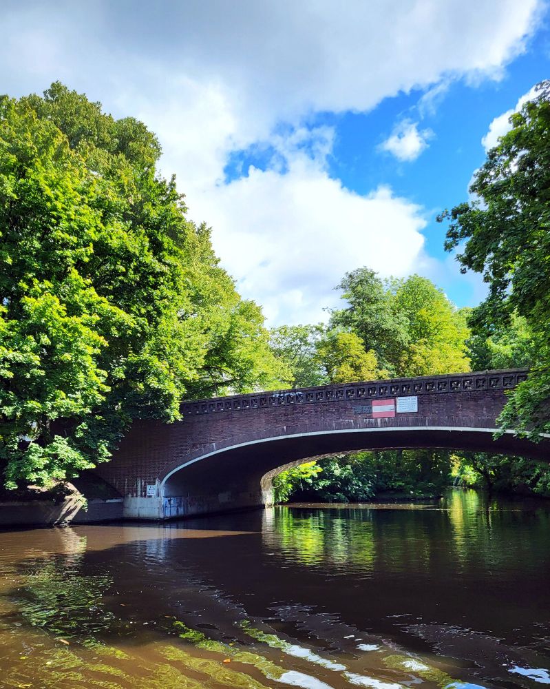 Blick auf die Alster. Eine große Steinbrücke. Links, rechts und Dahinter sind viele Büsche und Bäume. Der Himmel ist bewölkt.