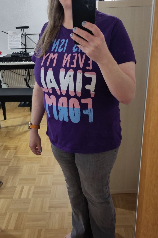 A mirror selfie of myself, with me wearing a pair of grey jeans and a purple teh shirt with the slogan “this isn't even my final form” written on it in the trans pride flag's colours: sky blue, pink and white.