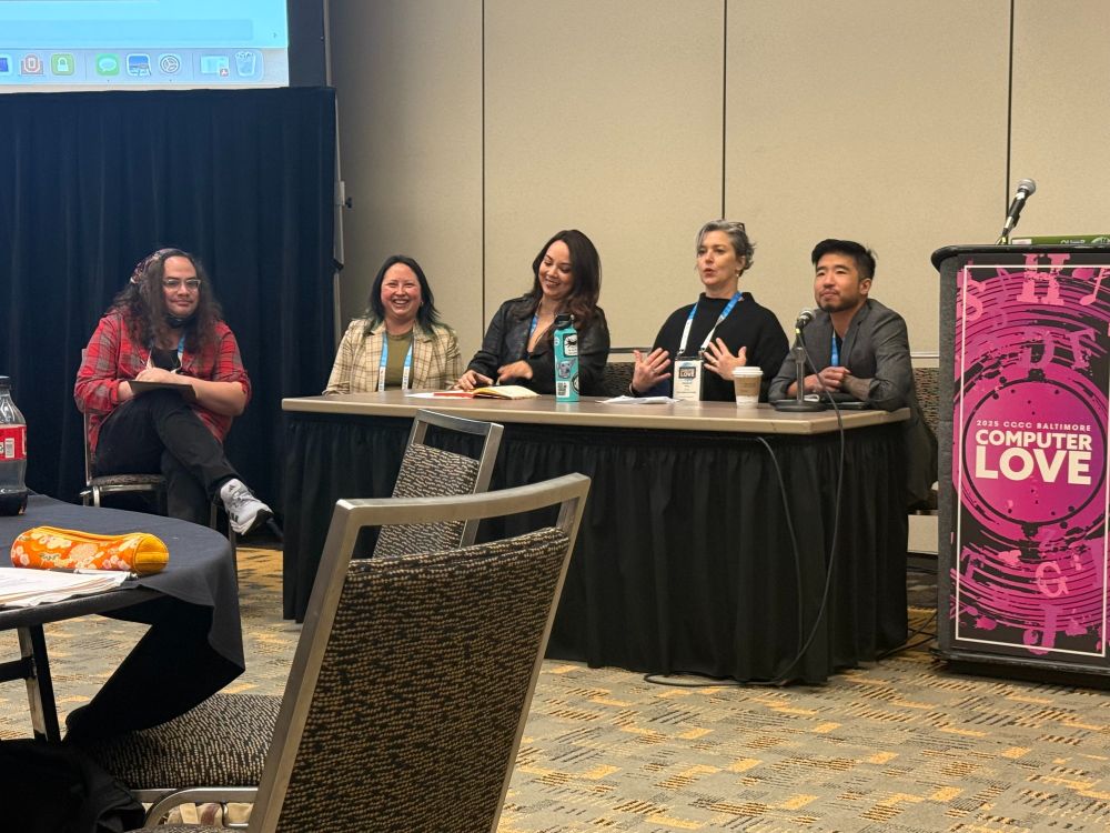 Five presenters of various races, ethnicities, genders, and outfits in red, black, gray, and cream sit behind a conference table responding to questions. Margaret is making a dramatic gesture toward her shoulders with both hands and the other folks on the roundtable are chuckling.