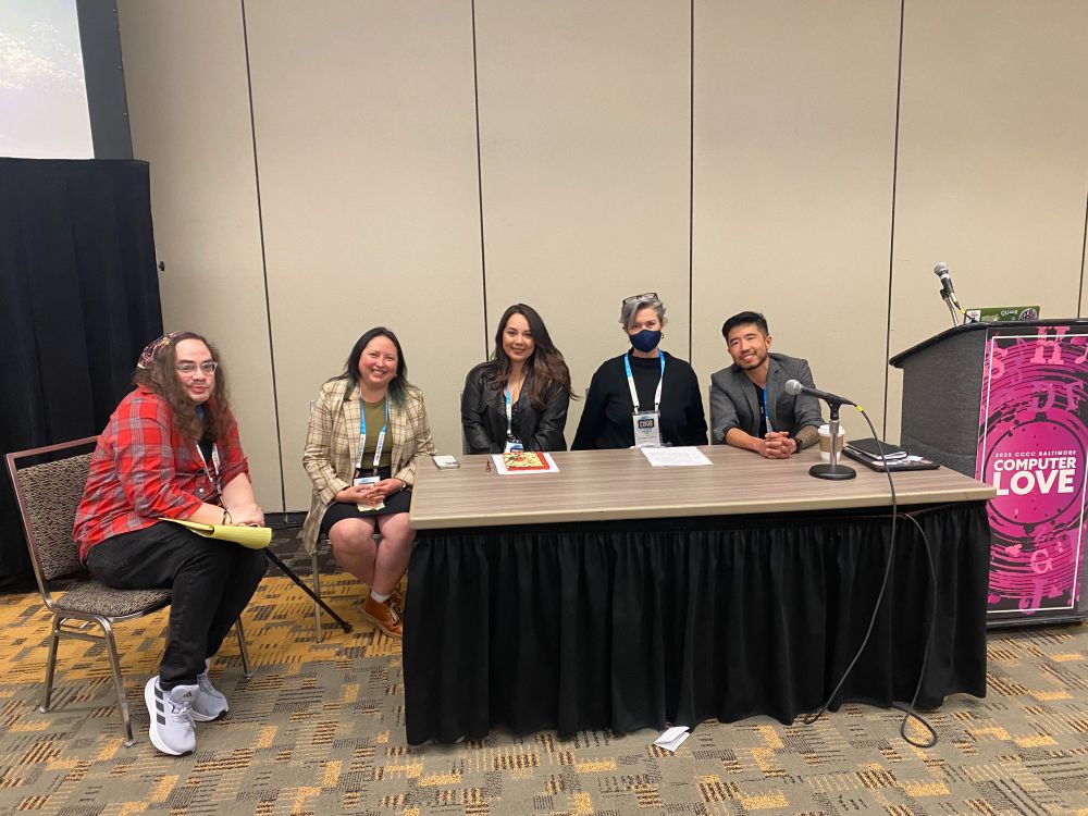 Five presenters of various races, ethnicities, genders, and outfits in red, black, gray, and cream sit behind a conference table smiling toward the camera.