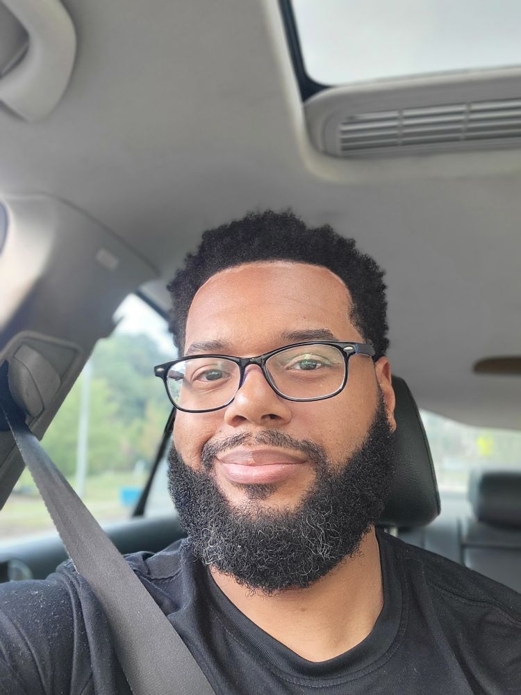 A somewhat handsome man wearing glasses and a black shirt smirking in a car