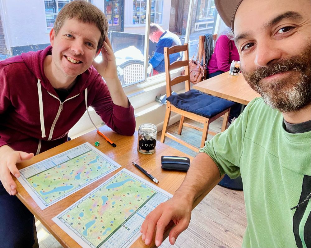 A selfie style photo showing two men playing a game at a coffee shop table. 