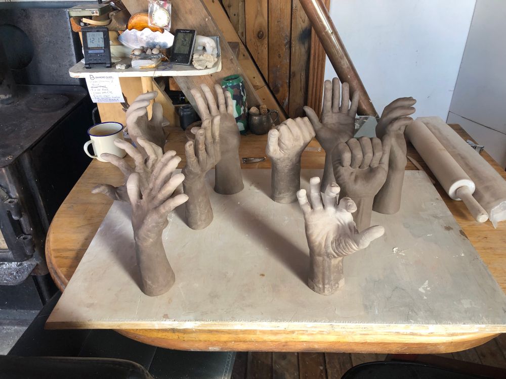 Ten clay sculptures of hands sit on a wooden table.  They randomly positioned in orientation, though all the sculptures start mid-way up the forearm.  They sit upright as though emerging from the table.  They are in different states of dryness, with colour variations from medium brown to tan.  Other objects sit on and around the table, showing a busy work space.