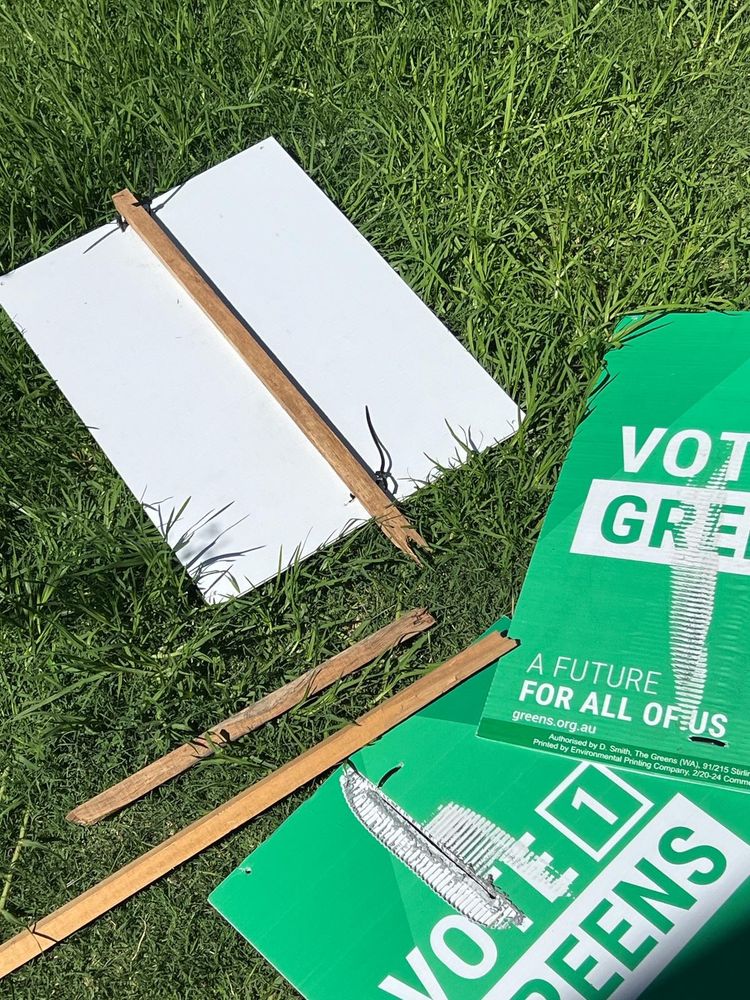 Three corflute signs laying in the green grass. One, top, is face down showing a white underside and broken wooden stake in the centre. The other two are face up with "Vote [1]" in white letters on a green background and Greens is green letters on a white box. Through the letters someone has cut into the sign damaging it.