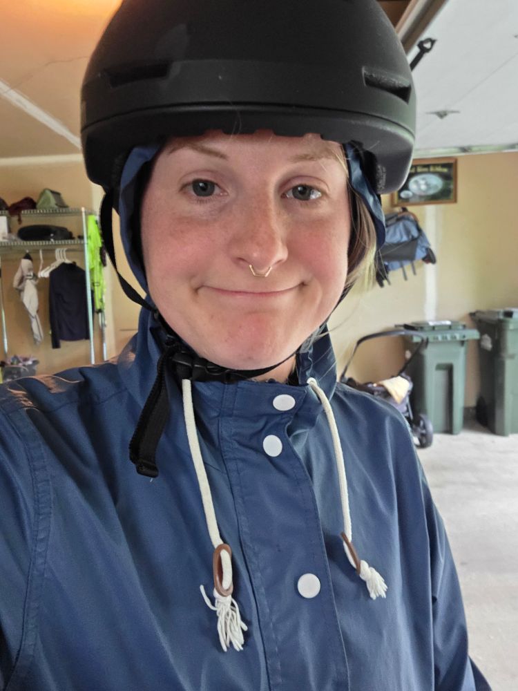 A photo of me standing in my garage wearing a blue rain jacket with the hood pulled up over my head. I have a black helmet on. I have a look of "welp here goes nothing" on my face.