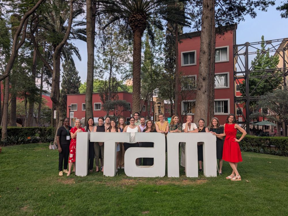 A group of scholars stand behind a sign that reads ITAM. There is green lawn to the front and sides of the sign and red buildings in the background.