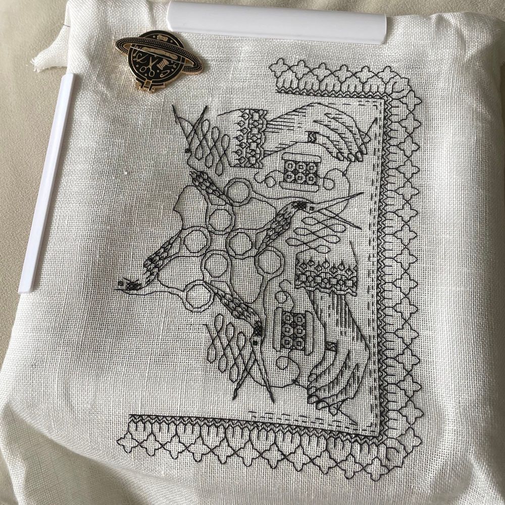 Blackwork embroidery on linen. It’s a circular design of a thread being unraveled from a bobbin, a sleeved hand sewing with the thread, & the thread being cut by a pair of bird scissors. It’s circular so that the process of sewing is repeated 4 times. There’s a square border surrounding everything. It’s unfinished but about 60% done. 