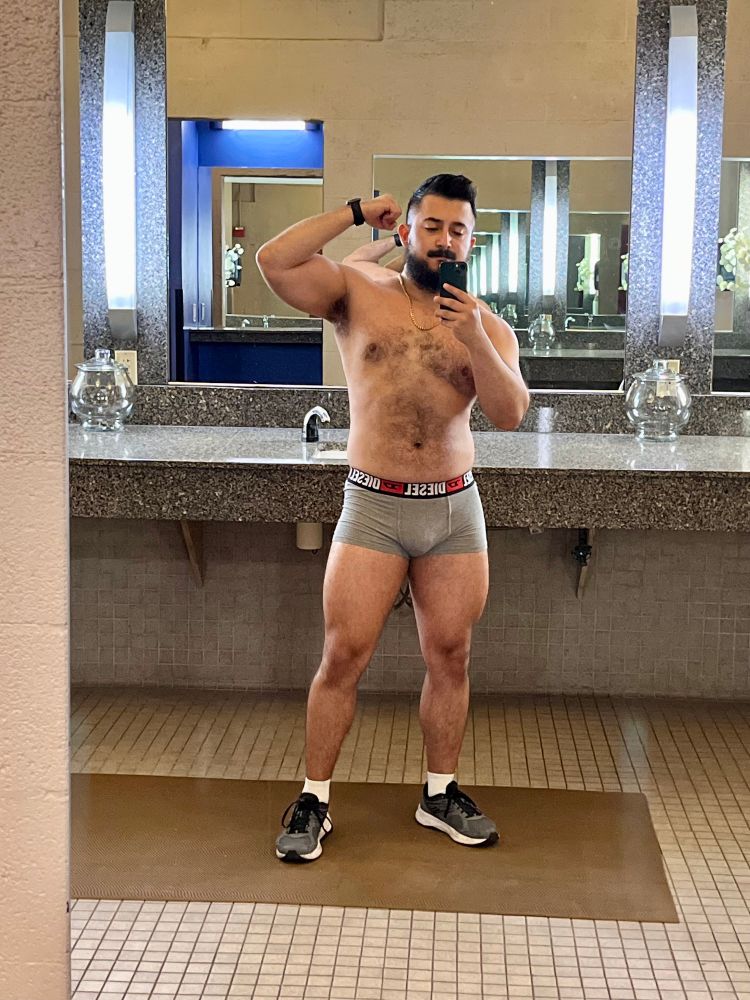 Progress pic at the gym against the mirror. I'm  shirtless, and wearing short trunks that show my leg muscles. I'm flexing my bicep.