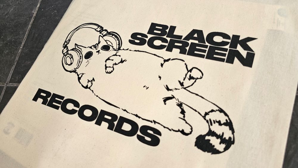 Black Screen Records fair trade cloth bag, showing a big cat illustration, laying on the side, wearing huge headphones, showing its blushy belly to the viewer while raising its head with huge, black pupils.