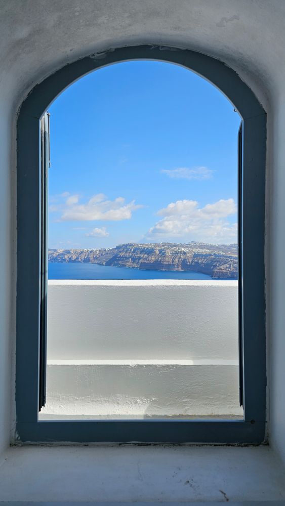 View from the bed: small window/balcony door, wide open with a view of part of the island of Santorini with sea, sunny weather, blue sky, and a few clouds.