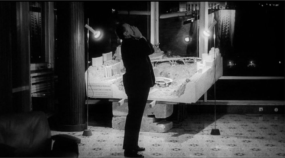 A man in a suit overs his hands over his face. He stands in an office room surrounded by glass windows a night. Lights shine over a large scale model of a real estate development building. 