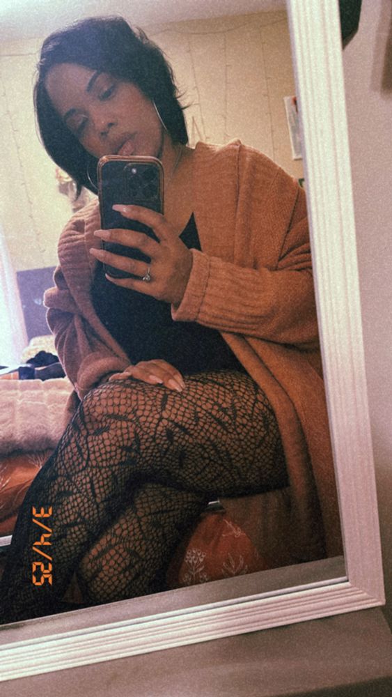 Black woman in pink cardigan and printed tights taking a selfie in a mirror with white trim 