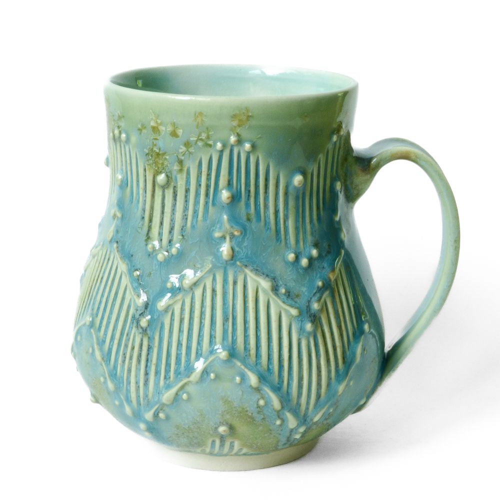 A mug with a zig zag geometric pattern created by carved lines and slip trailing. It has a turquoise crystalline glaze with more light blue and more green areas and green crystals 