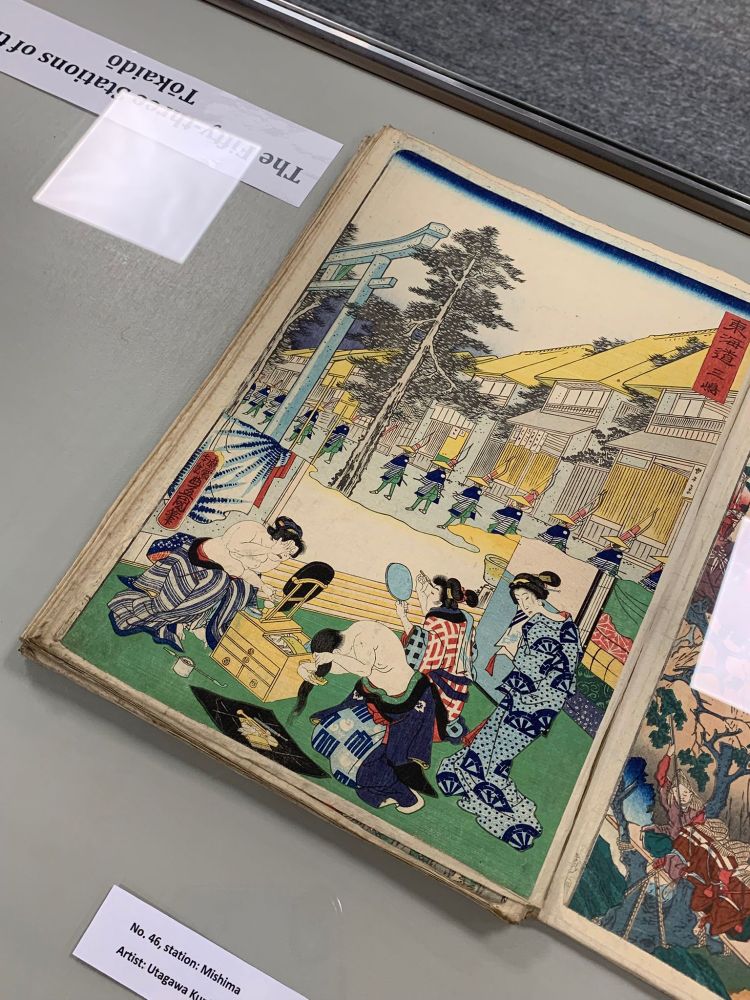 A fragile looking book sits under glass. It shows a full colour print of 4 geisha's dressing for the day whilst a row of soldiers wait in the courtyard outside.