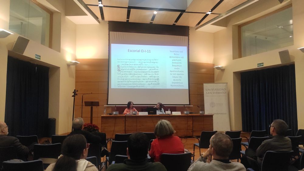 En la imagen se ve una sala dé ponencias. Al fondo se ve la pantalla con una imagen proyectada y dos personas sentadas de cara al público, que está de espaldas a la foto