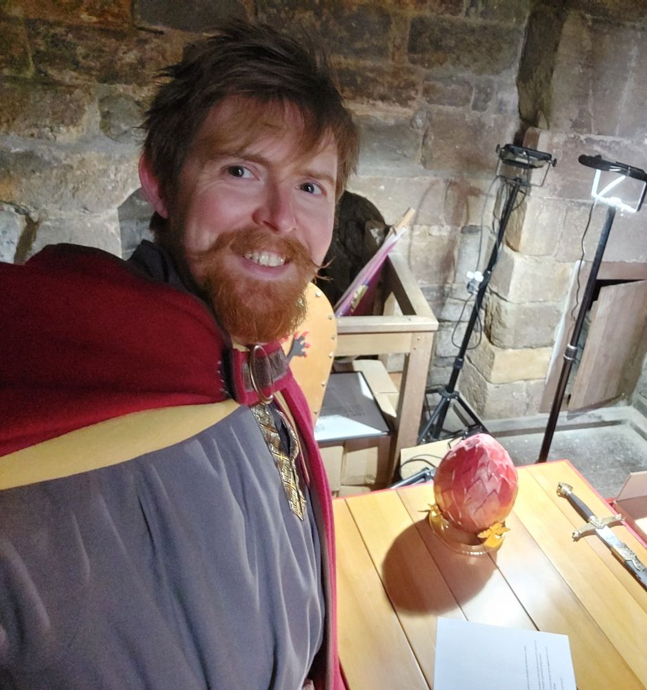 A guy in vaguely medieval costume standing by a table with a red paper-mache egg and sword on it