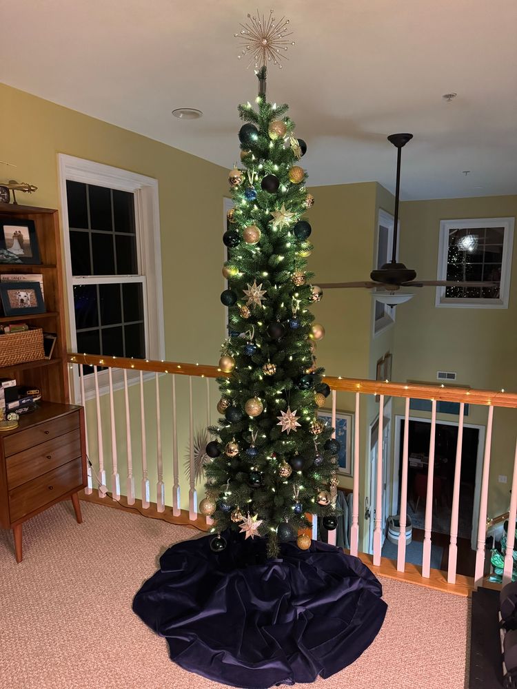 A tall skinny holiday tree decked in blue balls and gold stars. Beneath it is a deep blue velvet tree skirt.