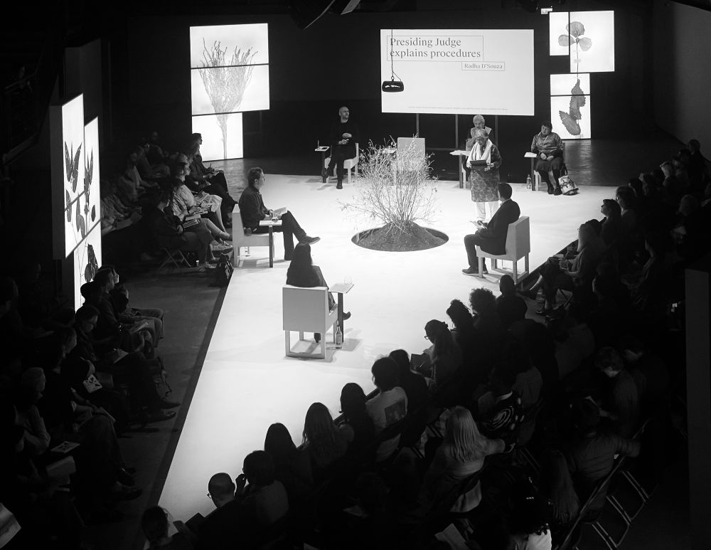 The Court room of the CICC at AmbikaP3, London. The court is located on a yellow wedge, modelled on El Lissitzky’s 1919 red wedge. There are human jurists around the two long sides of the wedge. An indigo plant comrade sits in attendance in a circle cut in the centre of the wedge, with large lightbox images of other subcontinental plant comrades exploited by the defendants. A video screen is placed behind the judge and other court officials to show evidence. Presiding judge Radha D’Souza is explaining how the court will proceed.