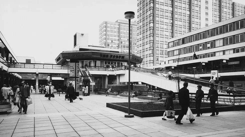 Black and white photo showing open-air shopping precinct on two levels, with a ramp and a staircase to the upper level, centred on a pub with an octagonal roof. The area is surrounded by tall office blocks and looks very 1970s.