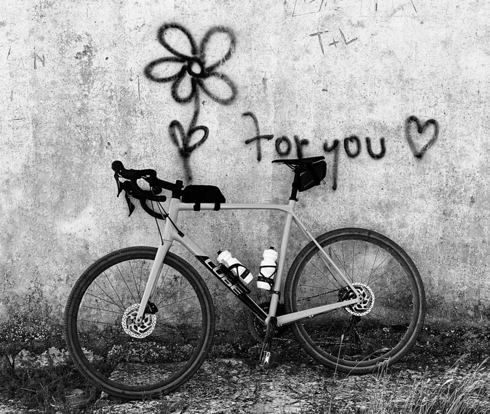 Ein Gravelbike lehnt an einer unverputzten Mauer. An der Mauer ein Graffiti, das aus einer Blume, den Worten „for you“ und einem Herz-Symbol besteht. Schwarz-weiß-Aufnahme.