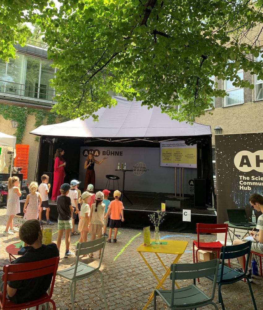Foto von der Veranstaltung, Kinder und bunte Stühle vor der Bühne. Auf der Bühne sind Lisa Rottenfußer und Moderatorin Andrea Lauterbach zu sehen, Lisa stellt den Kindern im Publikum eine Frage zu KI in der Wissenschaft.
