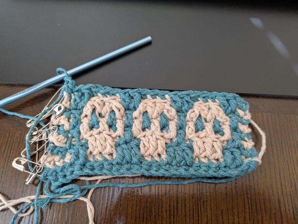 A small crochet project, about 4 inches by 2 inches. It's a skull pattern done in tan and blue green cotton yarn. There are safety pins lining the left side where the yarn should have been cut, but wasn't 