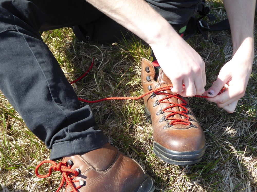 An actor lacing up walking boots viewed from above with the actor sitting and facing towards the camera. We can only see the boots, trousers, hands, and forearms of the actor. The boots are brown with red laces that have little flecks of black in them, the trousers are black cargo-style and the arms are bare - one thumb has the remains of some black nail polish on.