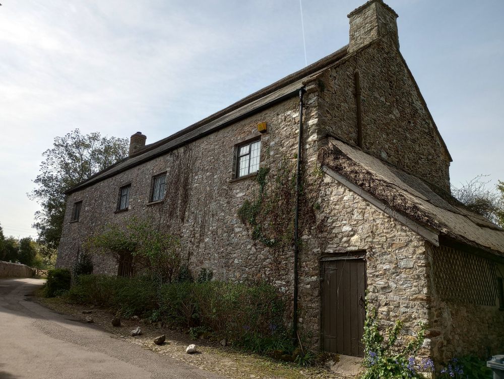 And old bare stone building with a tiled roof (although with a thatched apex). It has a cobbled path just in front of it which used to be part of a yard that has been tarmaced over to make the road. The main house is rectangular with small windows and a doorway about half-way along the ground floor. On the gable-end nearest the camera, a stone-built lean-to with thatched roof is a low doorway into the end of it. Small bushes grow against the walls, and a larger rose climbs over the main doorway, although nothing is in serious leaf yet.