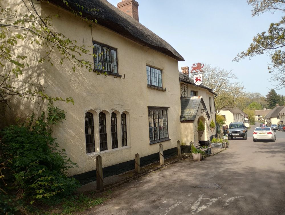A village street, concentrating on a public house. It has a hanging sign on it with a red lion passent depicted facing left, while above that, mounted on the same bar, is a cutout of a red lion passent, facing right.
The pub (and all the visible buildings) is thatched, painted a creamy yellow and clearly very old. The windows nearest the camera on the ground floor are a set of four, shallow arched & mullioned - the others a multi-paned using small glass squares of about 10cm. An arched entry porch is under the sign, about half-way down the building with two benches for seating on either side.
