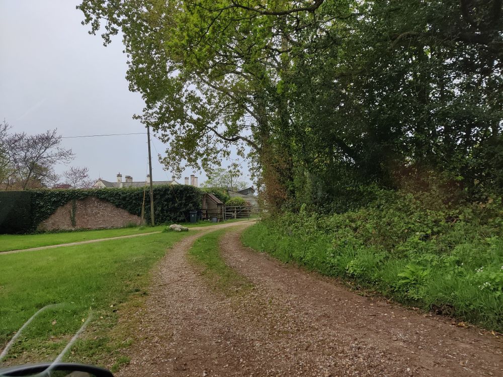 An unmetaled track leading around to a yard gateway in an extremely old wall, covered in ivy. Just over the wall, the roof apexes and chimneys of a large, old house can be seen.