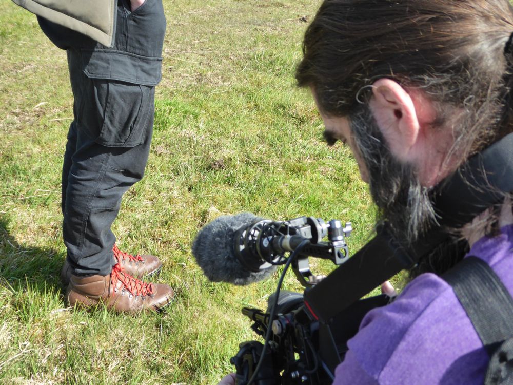 An actor and cameraman in the process of setting up an outdoor shot on short, rough grass. We can only see the actor from the waist down and they're wearing brown walking boots with red laces and black cargo trousers.
The cameraman (Me) is facing away from the shot and we're looking over his shoulder at the camera on a thick neckstrap with a shotgun mic mounted above it. The camera is pointed at the walking boots.