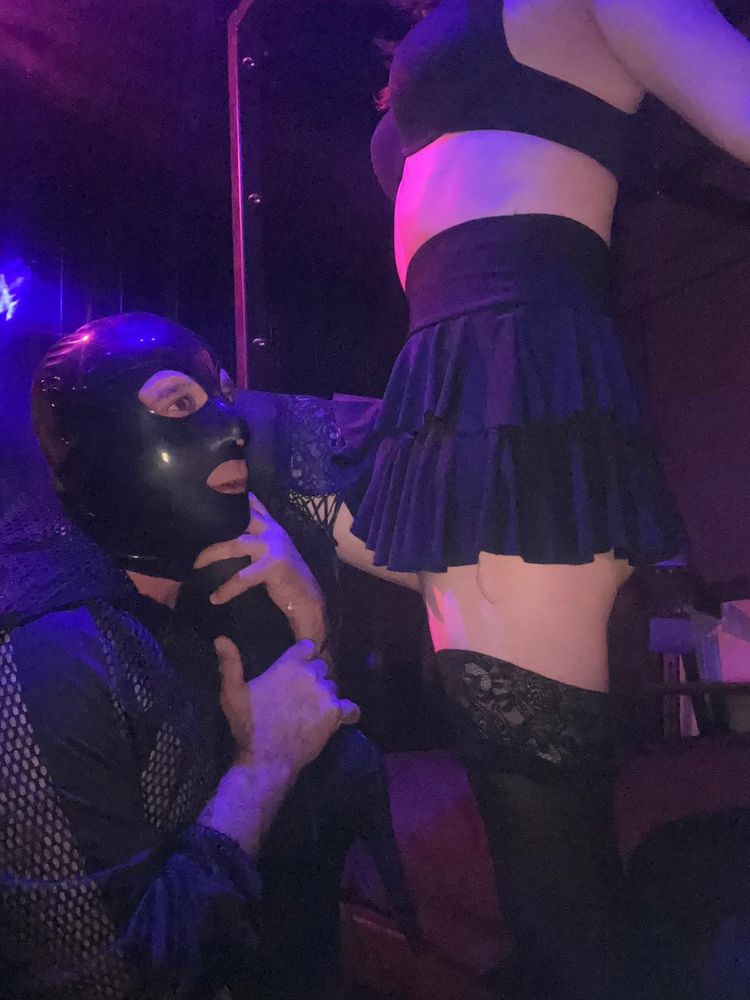 Trans Princess Jezebel dressed in black hold up stockings, frilly black skirt, bra and high heels stands in a dark dungeon space bathed in red light with a masked sub on his knees in front of her. She has her knee dominantly rested on his shoulder as he clutches on to her ankle and calf, peering up at her loyally.